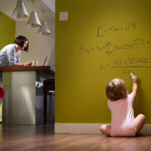 A toddler sitting on the floor works out complex mathematical equations on the wall while her mother nearby is oblivious